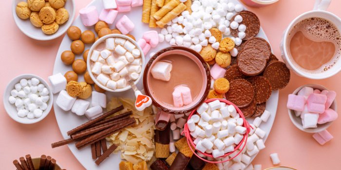 hot chocolate, marshmallows, chocolates and cookies charcuterie board on pink background closeup view, horizontal orientation